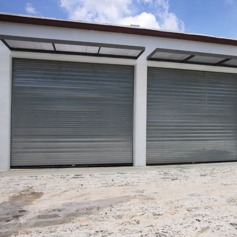 Two large grey metal rolling commercial garage doors installed on a white building with protective overhead grates, illustrating high-use industrial systems where safety and predictability are critical.