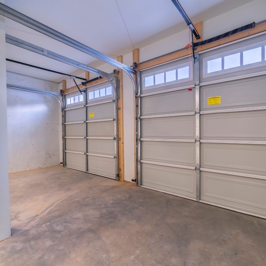Interior of a large residential garage in Greer showing two closed, grey sectional doors with windows, highlighting the heavy track and pulley systems that become unstable when off-track.
