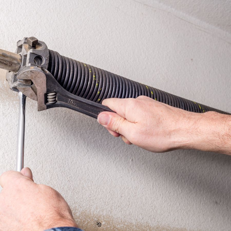 An close-up of a person's hands using an adjustable wrench and a metal rod to attempt a DIY garage door spring repair, illustrating the high-tension components that can cause sudden injuries or door failure if handled without professional tools and experience.