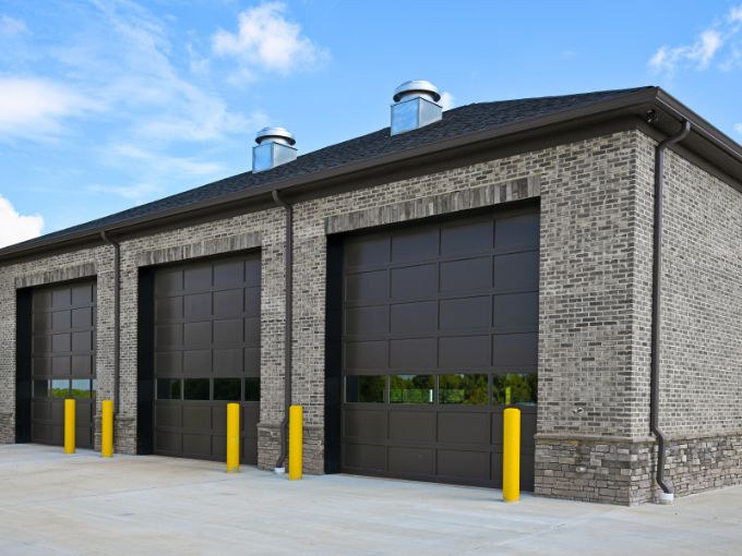 Three large dark-brown industrial sectional garage doors installed on a grey brick commercial building in Greer, SC, with yellow safety bollards positioned at the entrance.