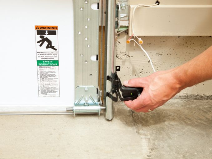 A close-up of a person's hand adjusting a black garage door safety sensor near the floor track of a white door in Greer, SC, with a visible "Warning" safety label on the door panel.